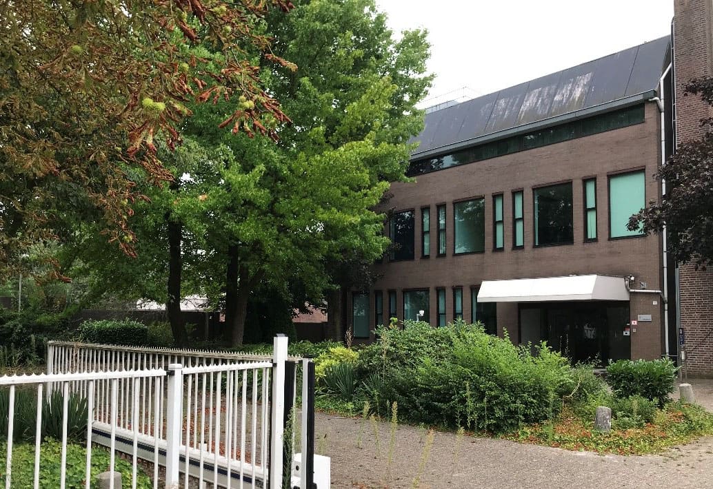 Een bakstenen gebouw met lichte luifel achter bomen en een wit hek.