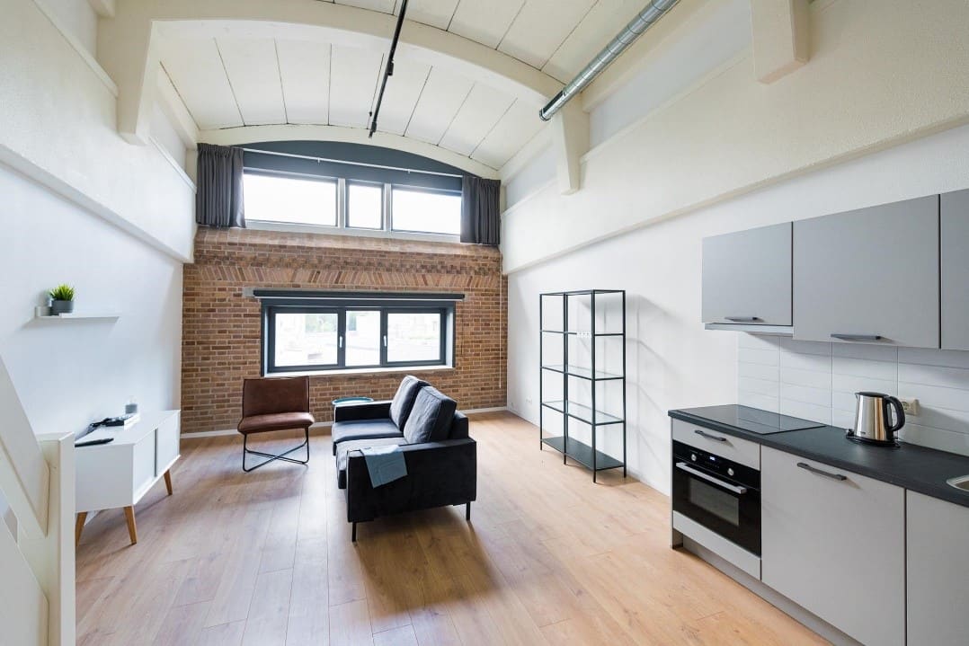 Een ingericht appartement met keuken, bank en een gebogen plafond onder een venster in de bakstenen gevel.