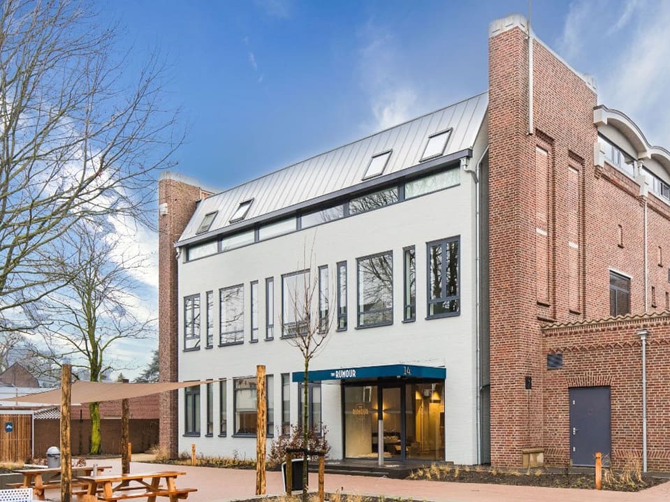 The Rumour na de verbouwing, met een wit gestucte gevel, de bakstenen kerkmuur en een terras op het voorplein.